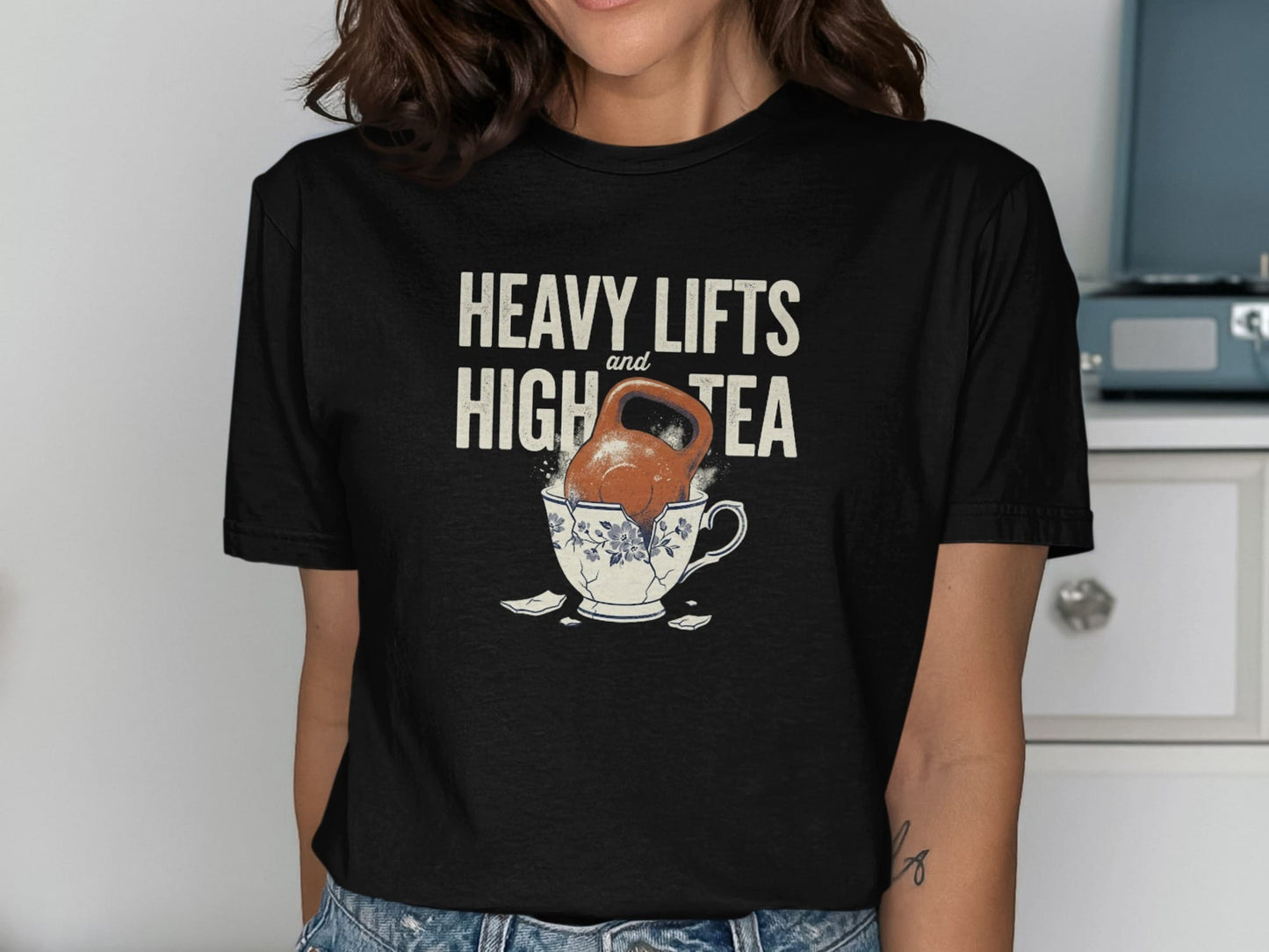 Black t-shirt with text "HEAVY LIFTS and HIGH TEA" over a kettlebell in a broken teacup.