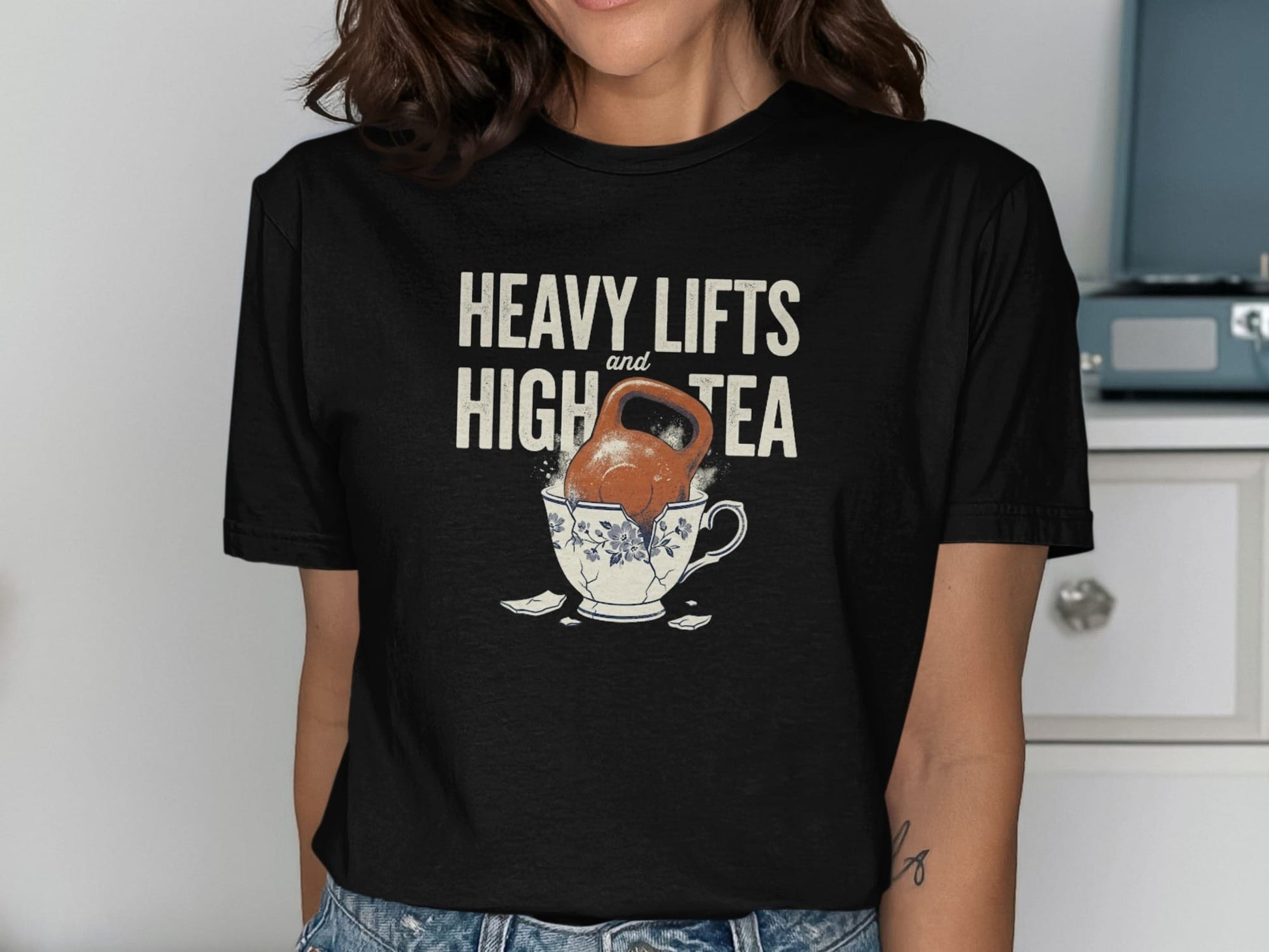Black t-shirt with text "HEAVY LIFTS and HIGH TEA" over a kettlebell in a broken teacup.