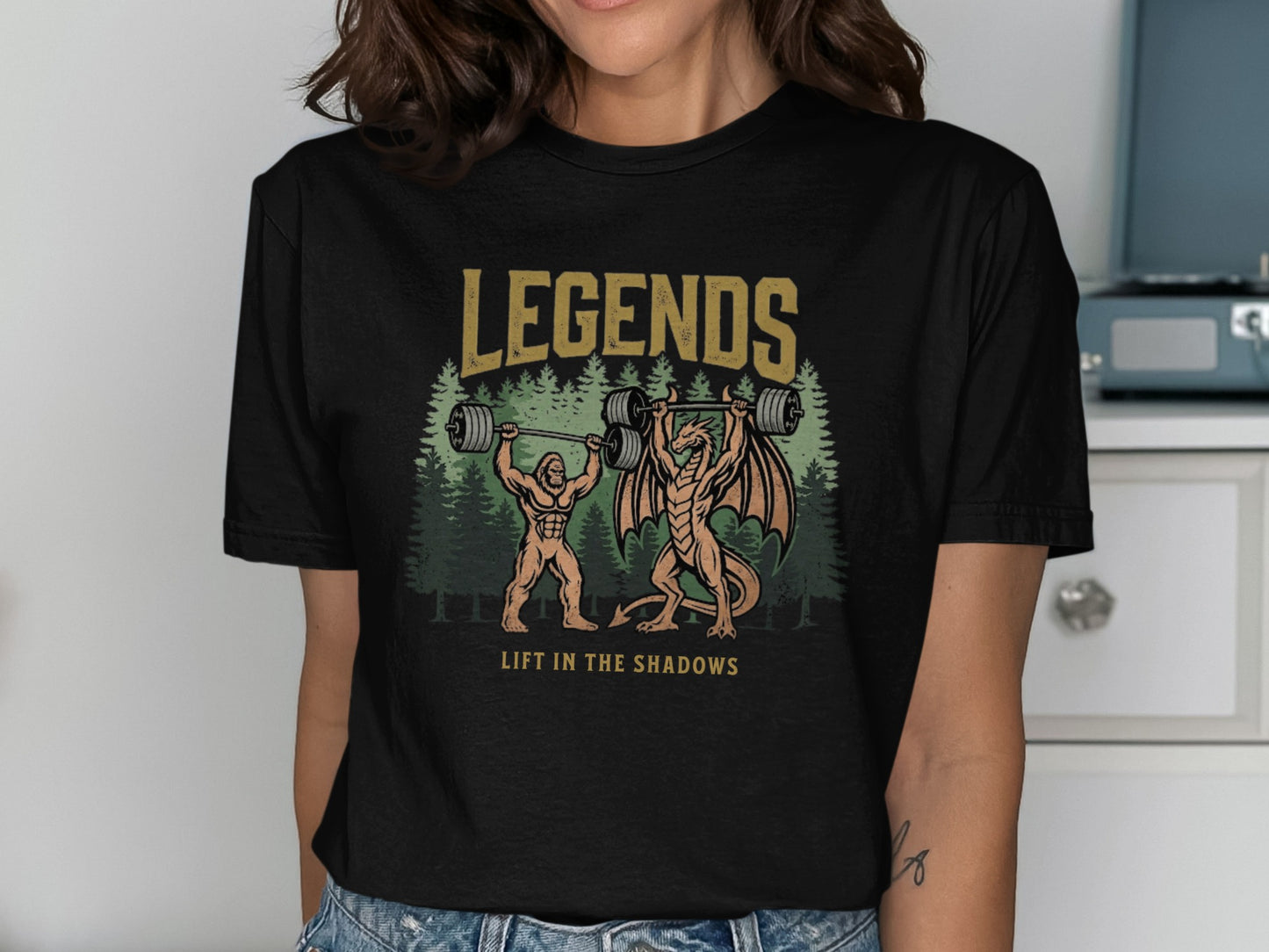 T-shirt graphic: Bigfoot and dragon lifting barbell in forest, text "LEGENDS LIFT IN THE SHADOWS".